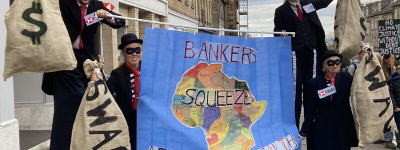 Four people, two on stilts, are dressed as bankers wearing robbers’ masks and holding big bags of swag. They are also holding a banner that shows an image of the continent of Africa that says ‘Bankers squeeze Africa’s wealth’. The banner is like a shower curtain so it can be gathered in the centre as if being squeezed.