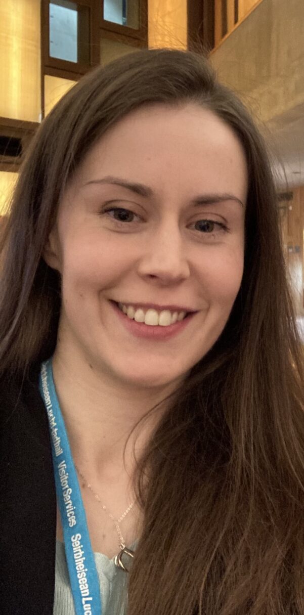 Headshot of Katelynne smiling at the camera. She is a white woman with long brown straight hair and is wearing a black suit jacket, a blue top and a blue lanyard. 